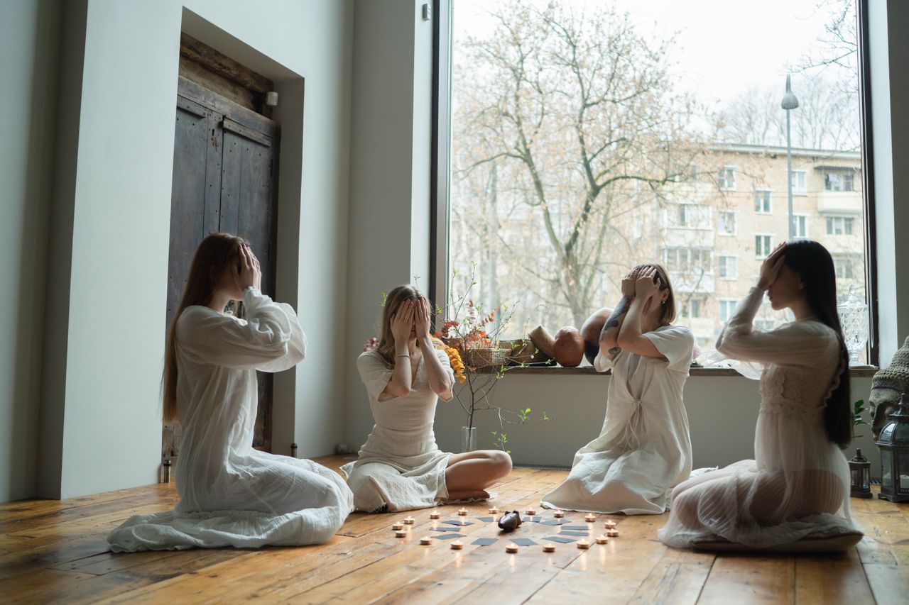 About Us Four women in white dresses engaged in a calming indoor ritual with candles and crystals.
